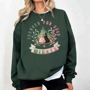 May include: Dark green sweatshirt with the embroidered text "CHANGED FOR GOOD" and "WICKED". The design features two figures in dresses, a castle, and floral accents in pink and green.