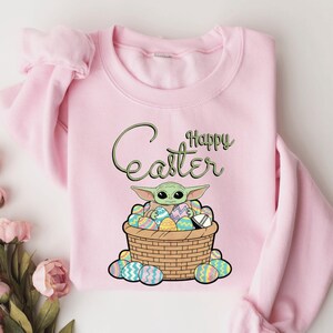 A pink sweatshirt with a graphic design featuring a young alien character, likely representing the character Baby Yoda from the Star Wars franchise, sitting in a basket filled with Easter eggs. The text "Happy Easter" is written above the image.