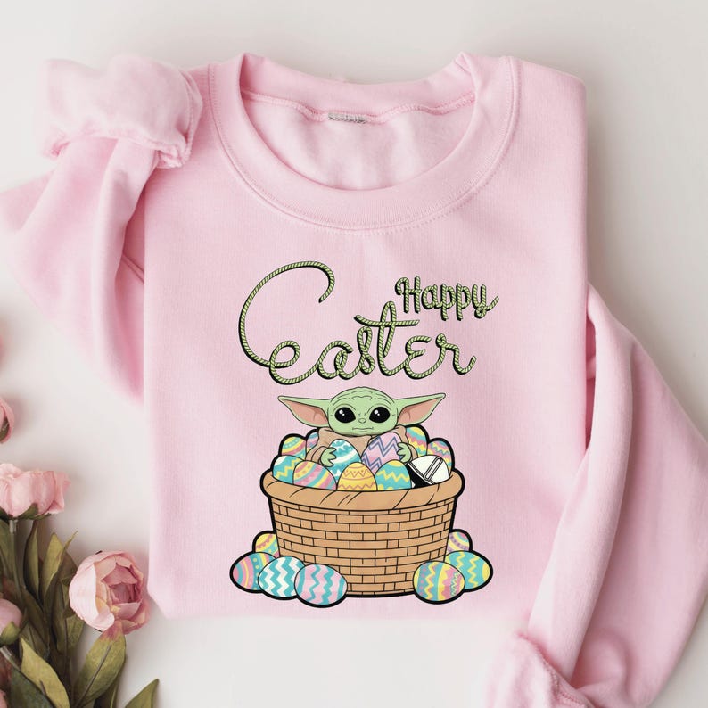 A pink sweatshirt with a graphic design featuring a young alien character, likely representing the character Baby Yoda from the Star Wars franchise, sitting in a basket filled with Easter eggs. The text "Happy Easter" is written above the image.
