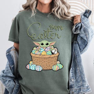 A person wearing a green t-shirt with a graphic of a young alien character, likely representing the character Baby Yoda from the Star Wars franchise, sitting in a basket filled with Easter eggs.
