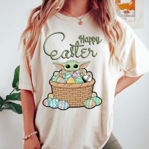 A woman wearing a white t-shirt with a graphic design of a young alien character, likely representing the character Baby Yoda from the Star Wars franchise, sitting in a basket filled with Easter eggs.