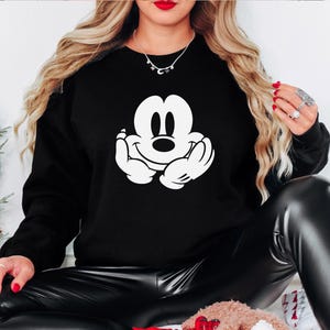 A woman wearing a black sweatshirt with a white Mickey Mouse logo is sitting on the floor.