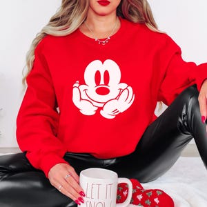 A woman wearing a red sweater with a white Mickey Mouse logo is sitting on the floor, holding a white mug with the text "LET IT SNOW" written on it.
