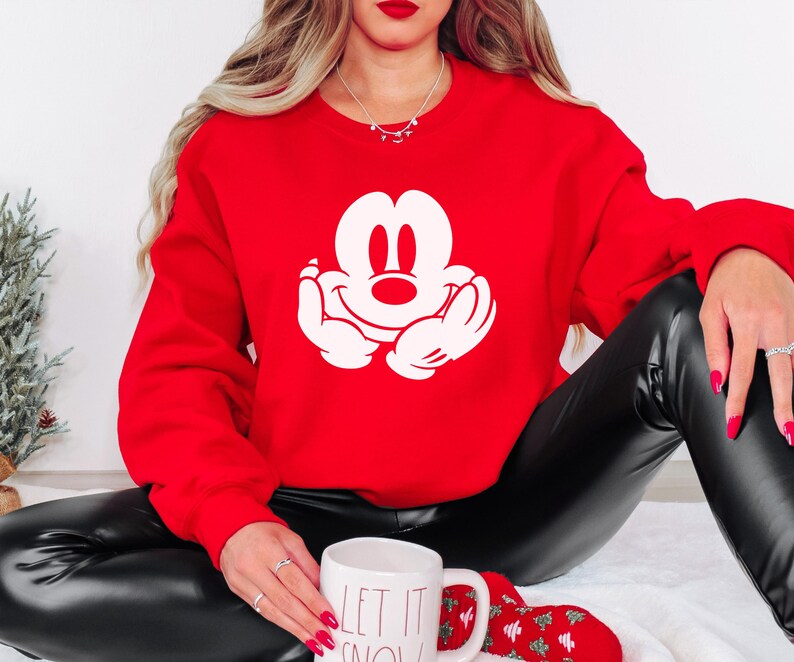 A woman wearing a red sweater with a white Mickey Mouse logo is sitting on the floor, holding a white mug with the text "LET IT SNOW" written on it.