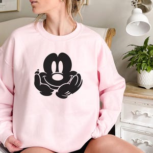 A woman wearing a pink sweatshirt with a black Mickey Mouse logo is sitting on a bed.