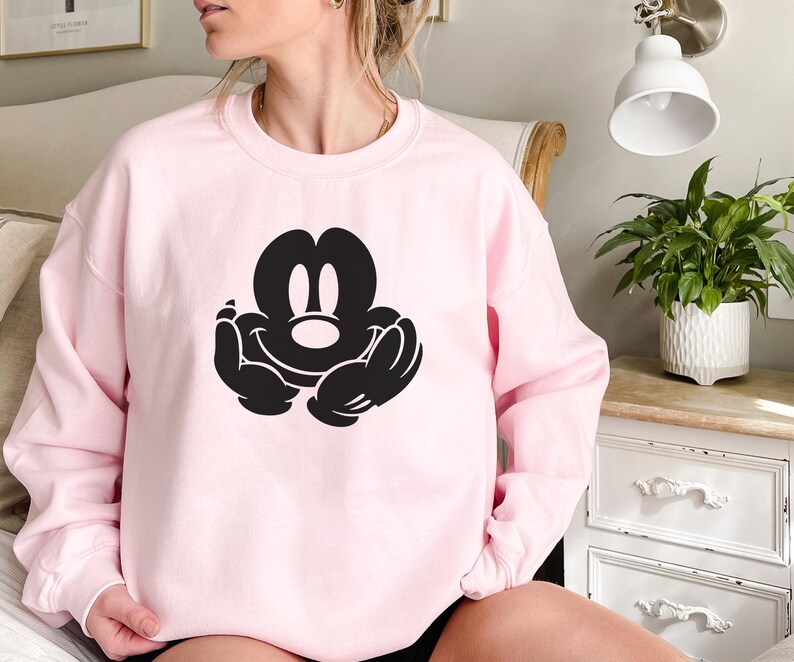 A woman wearing a pink sweatshirt with a black Mickey Mouse logo is sitting on a bed.
