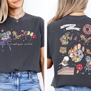 a woman wearing a gray t-shirt with a colorful graphic design on the back. The design includes cartoon characters such as a dog, a cat, and other animals, along with various objects like a donut, a hat, and a backpack. The text on the t-shirt reads "You can do anything you wish to be" and "The dog donut".