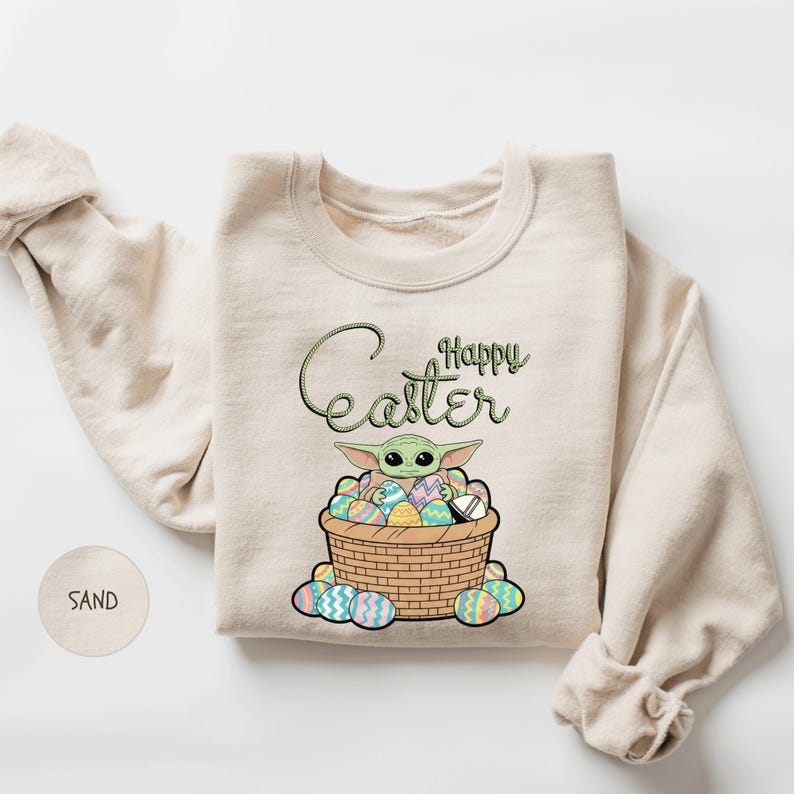 A beige sweater with a graphic design featuring a basket filled with Easter eggs and a small alien character.