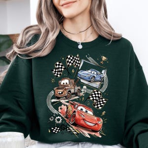 A woman wearing a green sweatshirt with a graphic design featuring three animated cars racing, including a red car, a blue car, and a brown truck.