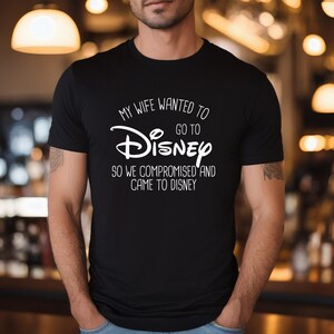 A man wearing a black t-shirt with white text that says "My wife wanted to go to Disney so we compromised and came to Disney".