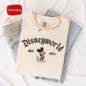 A white t-shirt with the text "Disneyworld EST. 1971" printed on it, along with a Mickey Mouse graphic, is laid out on a white surface.