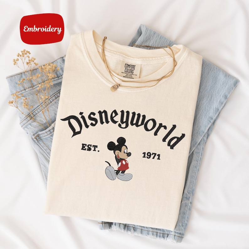 A white t-shirt with the text "Disneyworld EST. 1971" printed on it, along with a Mickey Mouse graphic, is laid out on a white surface.