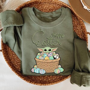 A green t-shirt with a graphic design of a young child, likely representing the character Baby Yoda from the Star Wars franchise, sitting in a basket filled with Easter eggs.