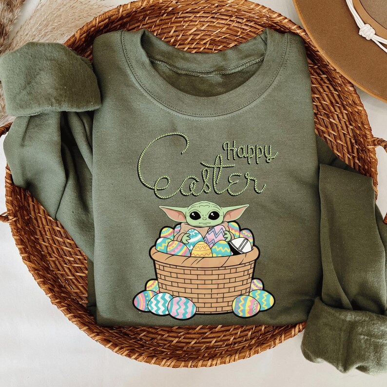 A green t-shirt with a graphic design of a young child, likely representing the character Baby Yoda from the Star Wars franchise, sitting in a basket filled with Easter eggs.