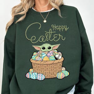 A green sweatshirt with a graphic design depicting a young alien character, likely representing the character Baby Yoda from the Star Wars franchise, sitting in a basket filled with Easter eggs. The text "Happy Easter" is written above the image.