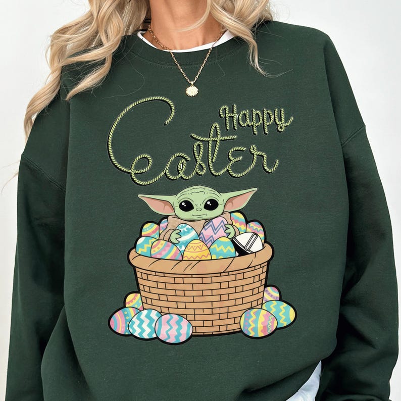 A green sweatshirt with a graphic design depicting a young alien character, likely representing the character Baby Yoda from the Star Wars franchise, sitting in a basket filled with Easter eggs. The text "Happy Easter" is written above the image.