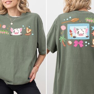 A woman wearing a green t-shirt with a cartoon pig design and various colorful elements, including a boat, flowers, and a bow.