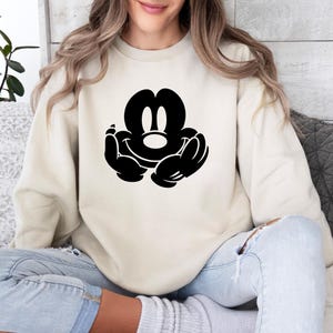 A woman wearing a white sweatshirt with a black Mickey Mouse logo is sitting on a couch.