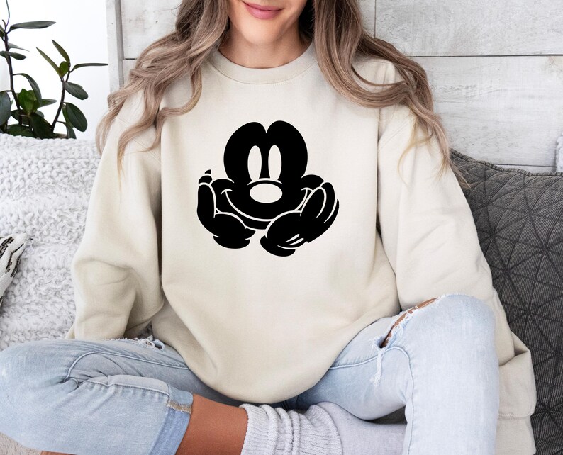 A woman wearing a white sweatshirt with a black Mickey Mouse logo is sitting on a couch.