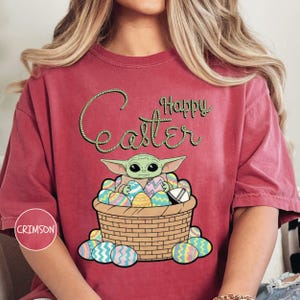 A person wearing a red t-shirt with a graphic design featuring a young alien character, likely representing the character Baby Yoda from the Star Wars franchise, sitting in a basket filled with Easter eggs.