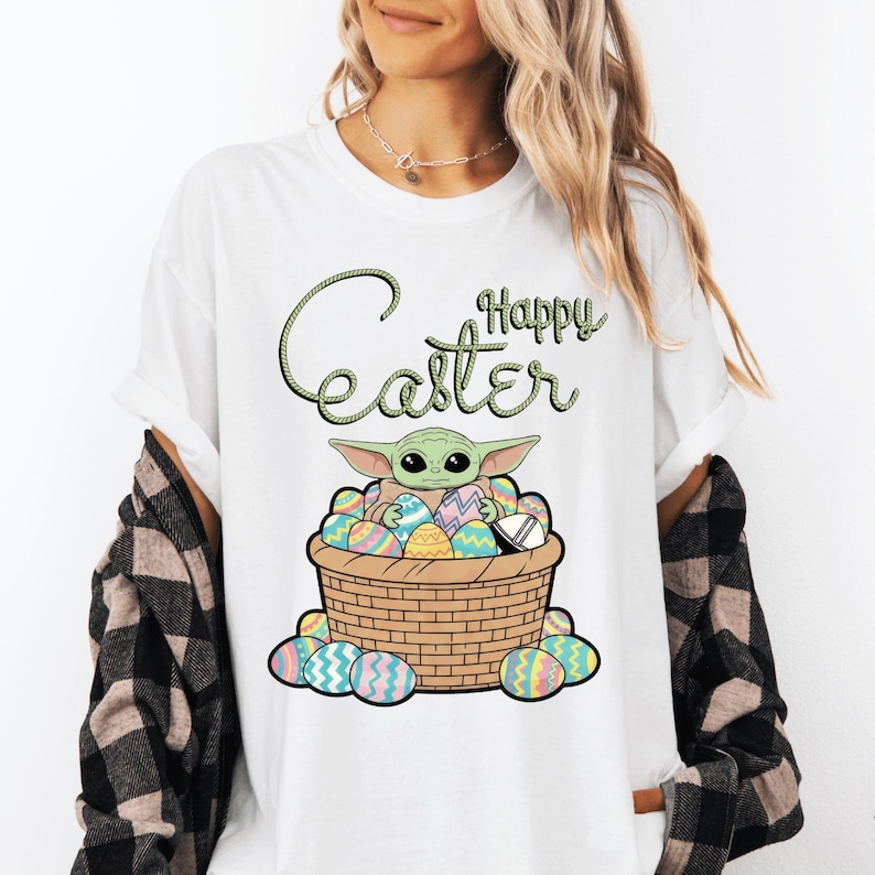 A woman wearing a white t-shirt with a graphic design of a young child, likely representing the character Baby Yoda from the Star Wars franchise, sitting in a basket filled with Easter eggs.