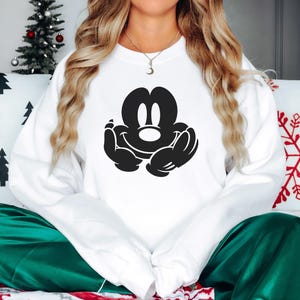 A person wearing a white sweatshirt with a black Mickey Mouse graphic is sitting on a bed with green pants and red and white pillows.