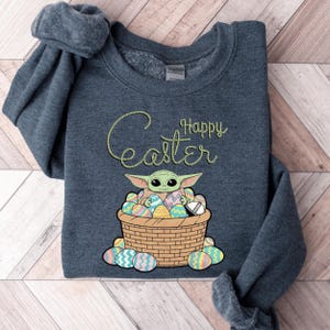 A gray sweatshirt with a graphic design featuring a young alien character, likely representing the character Baby Yoda from the Star Wars franchise, sitting in a basket filled with Easter eggs. The text "Happy Easter" is written above the character.