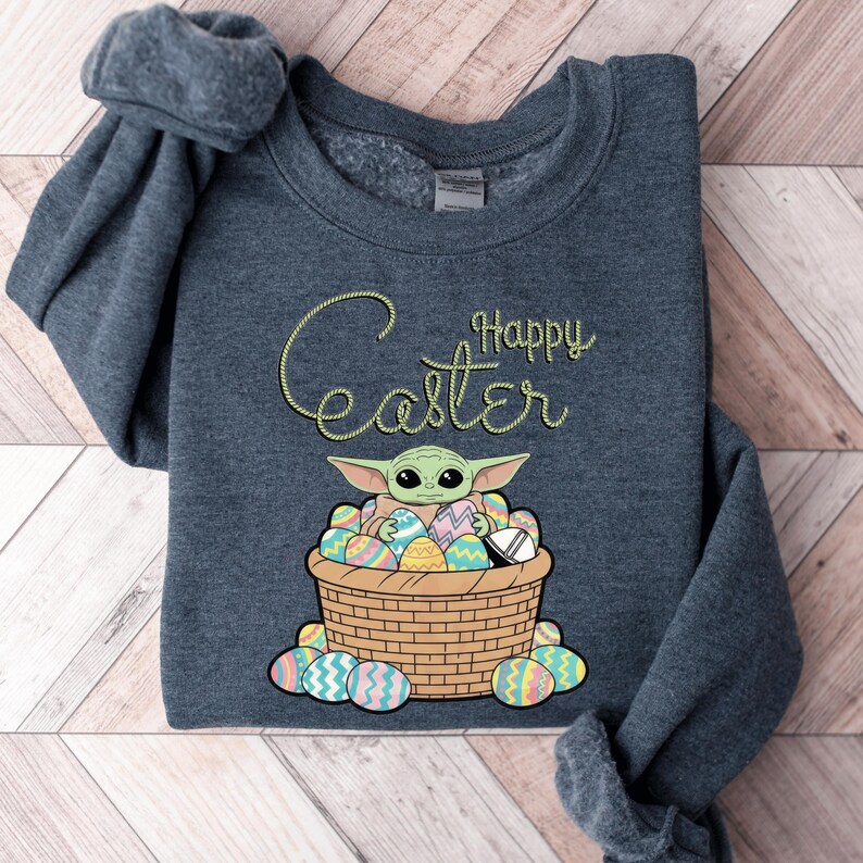 A gray sweatshirt with a graphic design featuring a young alien character, likely representing the character Baby Yoda from the Star Wars franchise, sitting in a basket filled with Easter eggs. The text "Happy Easter" is written above the character.