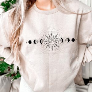 May include: Beige sweatshirt with a black and white celestial design. The graphic features a sunburst, moon phases, and stars. The sweatshirt is a neutral colour and has a crew neck.