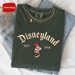 A green t-shirt with the text "Disneyland EST. 1955" and a Minnie Mouse embroidered on it, along with a yellow chain.