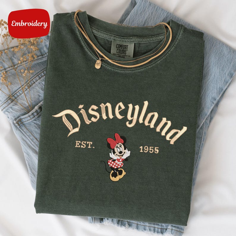 A green t-shirt with the text "Disneyland EST. 1955" and a Minnie Mouse embroidered on it, along with a yellow chain.