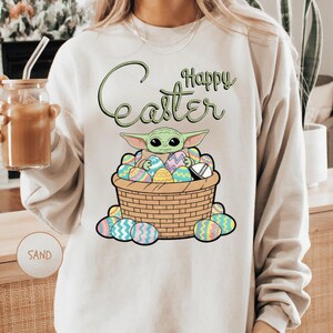 A person wearing a white sweater with a graphic design featuring a young alien character surrounded by colorful Easter eggs.