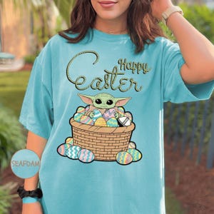 A woman wearing a teal t-shirt with a graphic of a young child, likely representing the character Baby Yoda from the Star Wars franchise, sitting in a basket filled with Easter eggs.