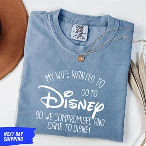 A blue t-shirt with white text that says "My wife wanted to go to Disney so we compromised and came to Disney" is displayed on a white surface.