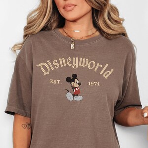 A woman wearing a brown t-shirt with the text "Disneyworld EST. 1971" and a Mickey Mouse graphic.