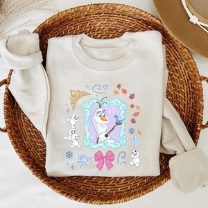 A white t-shirt with a graphic design featuring Olaf, the main character from the Disney movie Frozen, along with other snowmen and snowflakes.