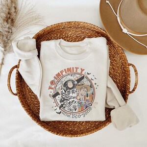 A white sweatshirt with a graphic design featuring a character from the Toy Story franchise, along with the text "To infinity and beyond" and "and Eco-yong". The sweatshirt is placed in a woven basket, and there is a hat visible in the background.