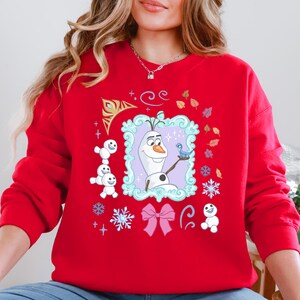 A woman wearing a red sweater with a graphic of Olaf, the main character from the Disney movie Frozen, surrounded by various snow-themed elements.