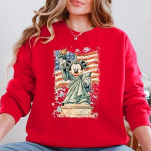 A woman wearing a red sweatshirt with a graphic of Mickey Mouse holding the torch of freedom in front of the Statue of Liberty.