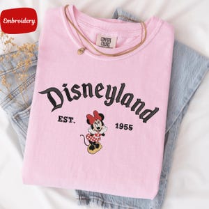 A pink t-shirt with the text "Disneyland EST. 1955" and a Minnie Mouse graphic, along with a necklace, is displayed on a bed.