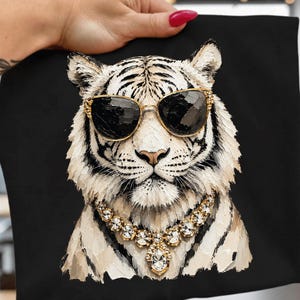 Könnte beinhalten: Ein schwarzes T-Shirt mit einem strukturierten Gemälde im Impasto-Stil, das einen weißen Tiger zeigt. Der Tiger trägt eine goldgerahmte Sonnenbrille und eine goldene Halskette mit klaren, facettierten Kristallen. Das Kunstwerk zeigt sichtbare Pinselstriche.