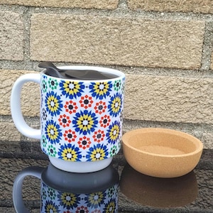 May include: A white ceramic mug with a colorful geometric pattern, a black spoon, and a small cork bowl. The mug's design features blue, yellow, red, and green shapes. The items are on a reflective surface.
