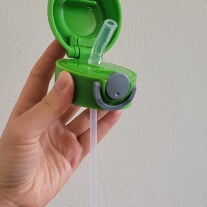 Puede incluir: Una tapa de botella de agua de pl&aacute;stico verde con una abertura abatible y una pajita transparente. La tapa tiene un asa gris y un peque&ntilde;o bot&oacute;n redondo gris. La pajita se extiende hacia abajo.
