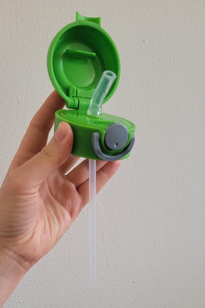 Puede incluir: Una tapa de botella de agua de pl&aacute;stico verde con una abertura abatible y una pajita transparente. La tapa tiene un asa gris y un peque&ntilde;o bot&oacute;n redondo gris. La pajita se extiende hacia abajo.