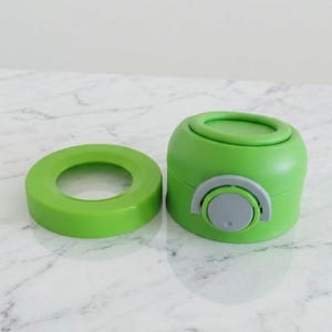 Puede incluir: Un juego de tapa y base de botella de agua de color verde brillante. La tapa es un anillo circular, y la base es un cilindro redondeado con un bot&oacute;n gris. El juego es de pl&aacute;stico.