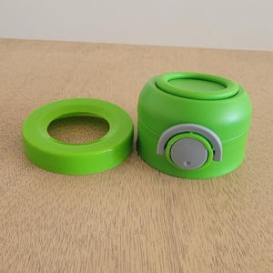 Puede incluir: Juego de tapa y tap&oacute;n verde para botella de agua. La tapa es una pieza plana y circular, y el tap&oacute;n es un cilindro redondeado con una palanca gris y blanca. El juego es de pl&aacute;stico.