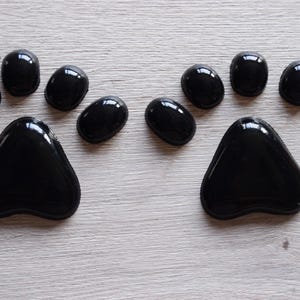 May include: Two sets of black paw prints are arranged on a light wood-grain surface. Each paw print consists of a large, triangular pad and four smaller, oval-shaped toe pads. The glossy black stones create a striking contrast.