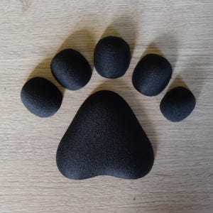 May include: A black paw print made of six textured, rounded shapes. The large pad is triangular, and the smaller toe pads are arranged above it. The paw print is set against a light brown wooden surface.