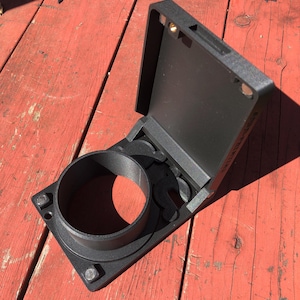 May include: A black, open electrical box with a circular opening and a hinged lid. The box is mounted on a square base with bolts. The lid is open, revealing the interior. The box is made of a matte black material.