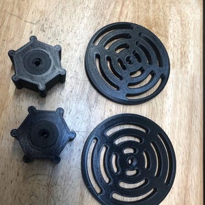 May include: Four black 3D-printed objects are displayed on a wooden surface. Two are hexagonal with a central circular feature, and two are circular with a series of concentric arcs. The objects appear to be components or parts.